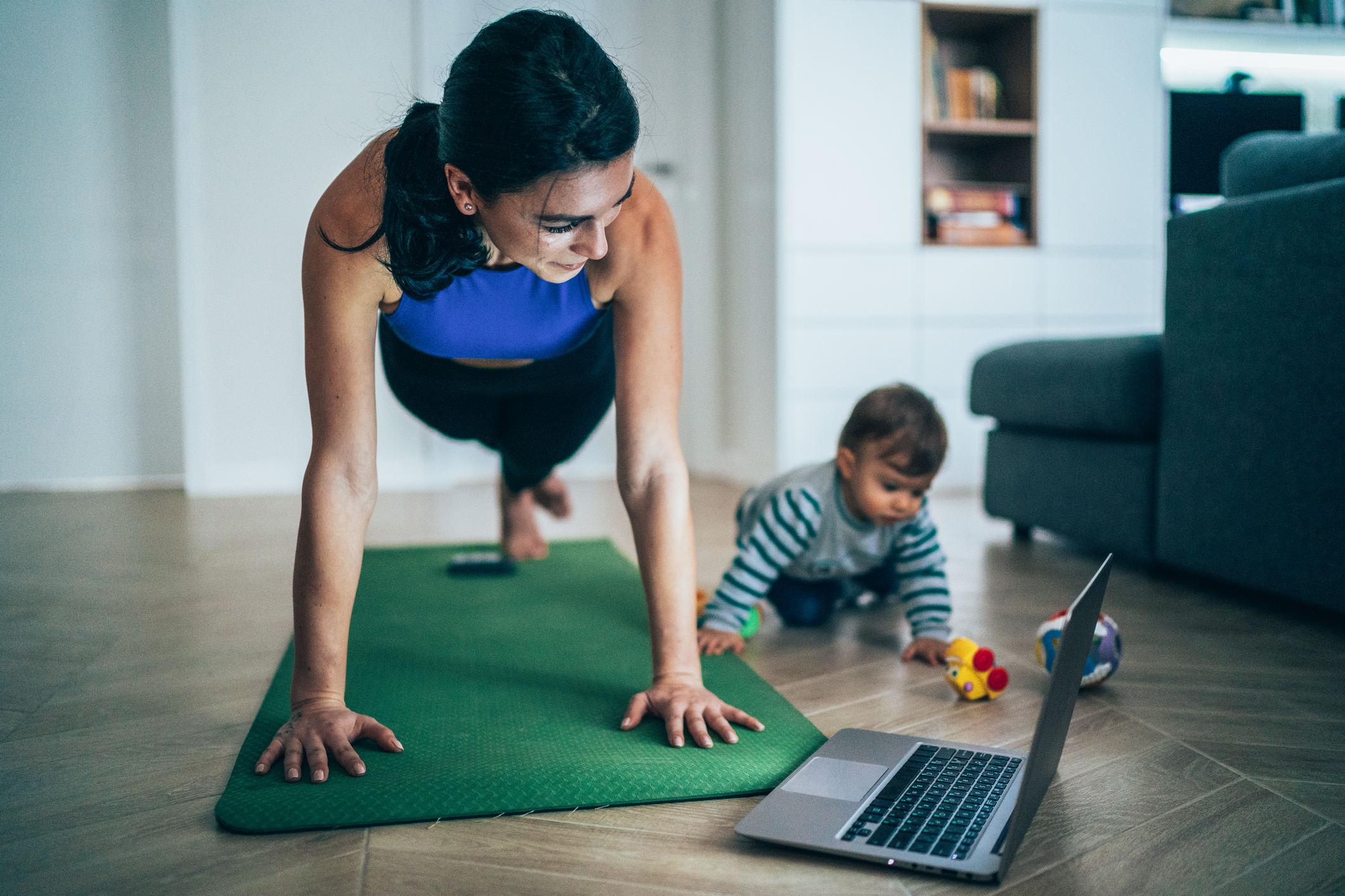 女性一边在笔记本电脑上观看锻炼视频，一边和她的孩子玩耍