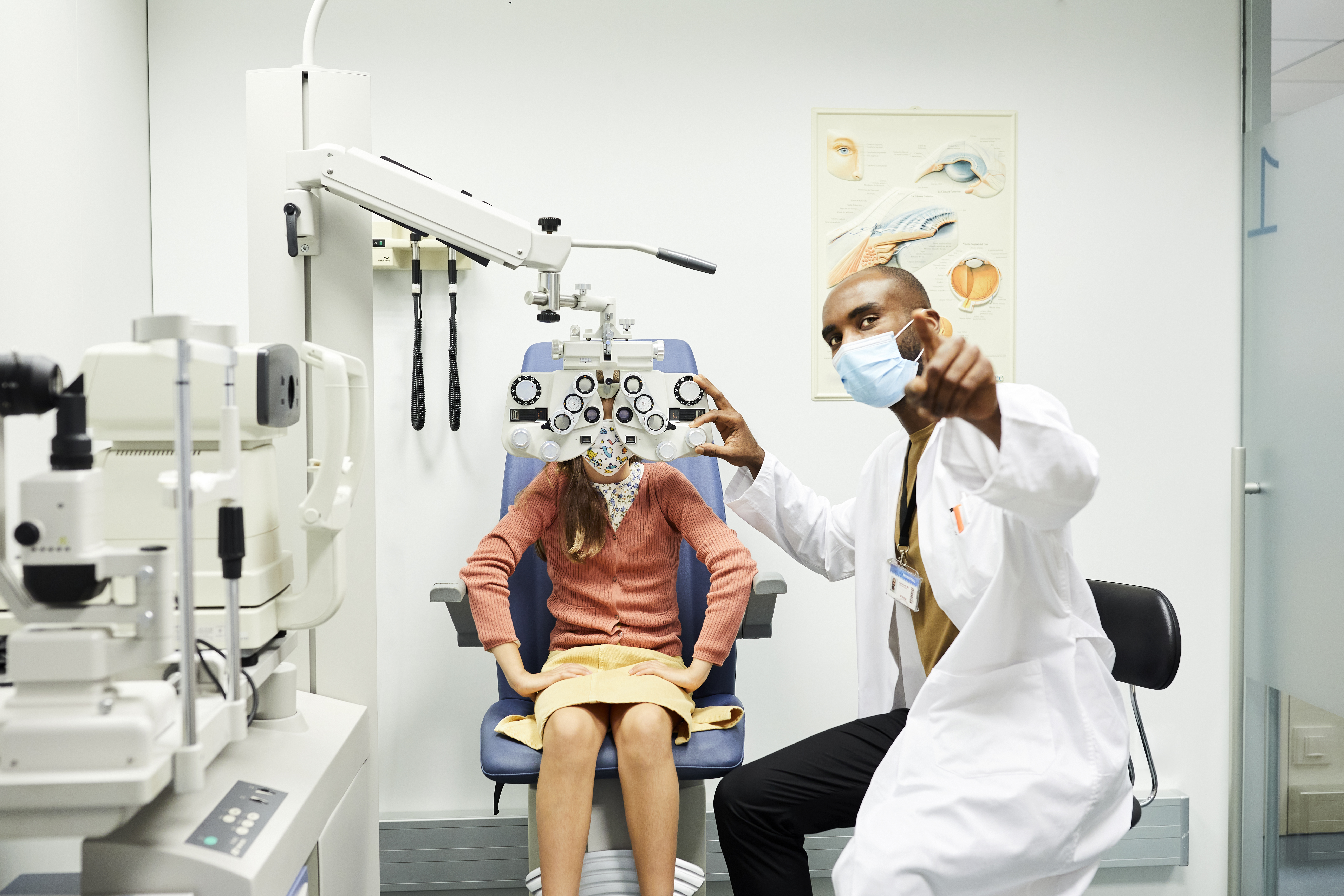眼科医生戴着防护面罩向女孩做手势。女童坐在医生诊所验光仪的后面。他们在 COVID-19 危机期间在医院接受眼科检查。