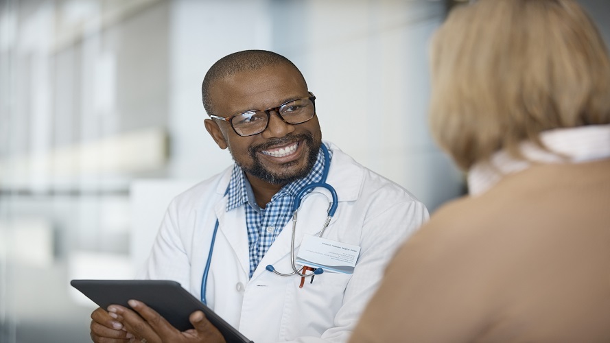 微笑的男医生拿着平板电脑，看着年长的女性。患者正在医院找医疗人员就诊。他穿着实验服，戴着眼镜。