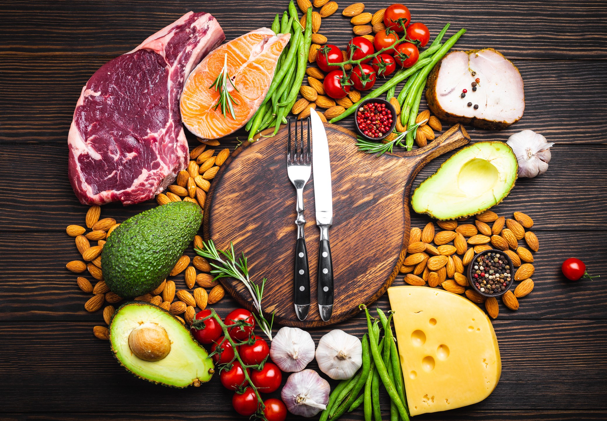 木制砧板上的刀叉，以及针对健康饮食概念和减肥的生酮低碳水化合物食材，顶视图。酮类食物：肉、鱼、鳄梨、奶酪、蔬菜、坚果