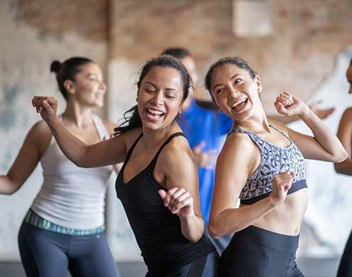 A small group of Hispanic men and women participate in a fitness dance class.  They are wearing comfortable athletic wear and have their arms p as they dance and move around the room with joyful expressions.